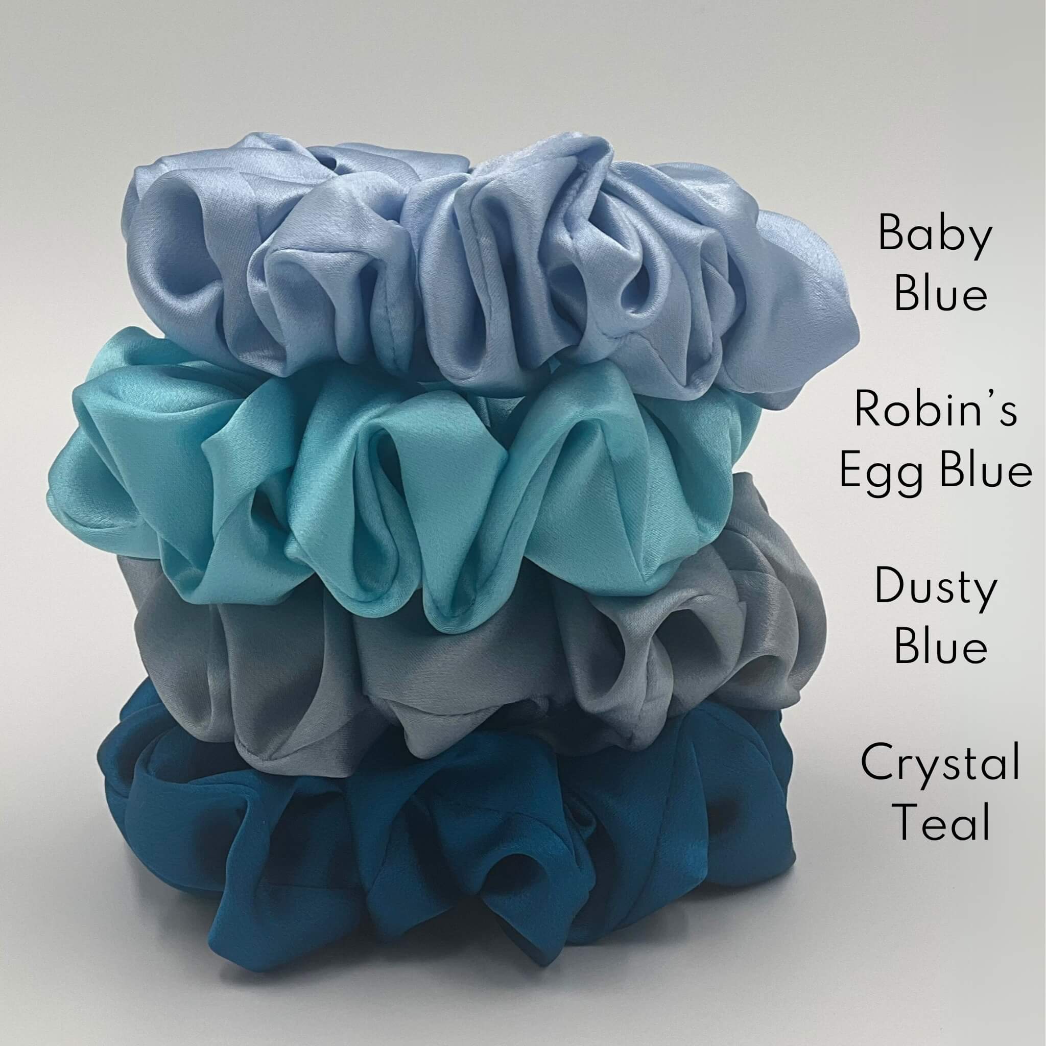 A pile of four different coloured blue silk scrunchies with their colour names next to them in black font "baby blue", "robin's egg blue", "dusty blue" and "crystal teal"