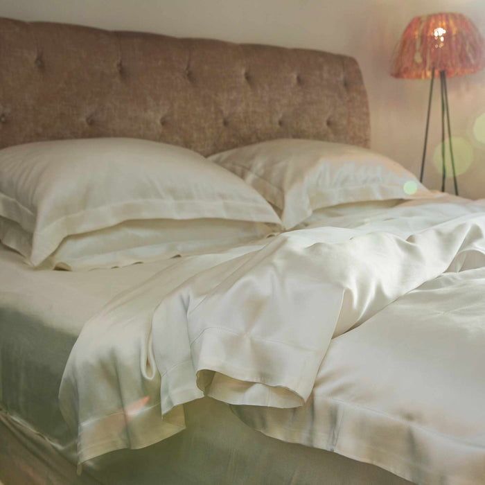 Neatly made bed with ivory silk bedding and pillows, illuminated by a lamp in a softly lit room.