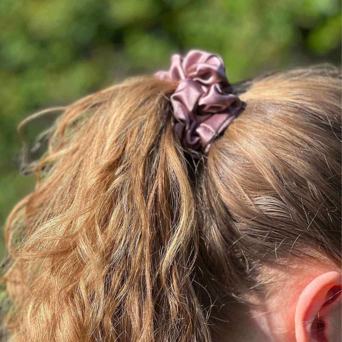 Person with a dusty pink mulberry silk scrunchie in their hair against a blurred green background