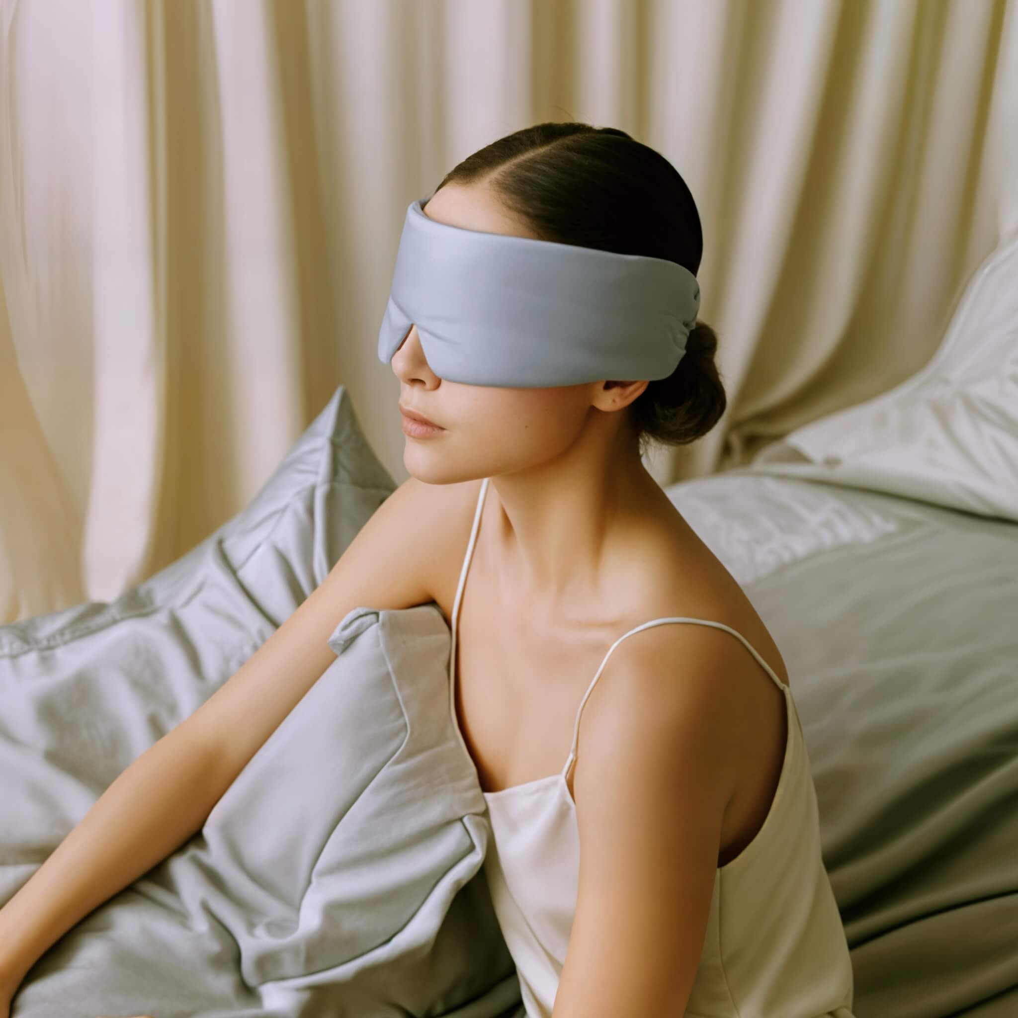 Woman wearing a light blue silk sleep mask in a bedroom setting