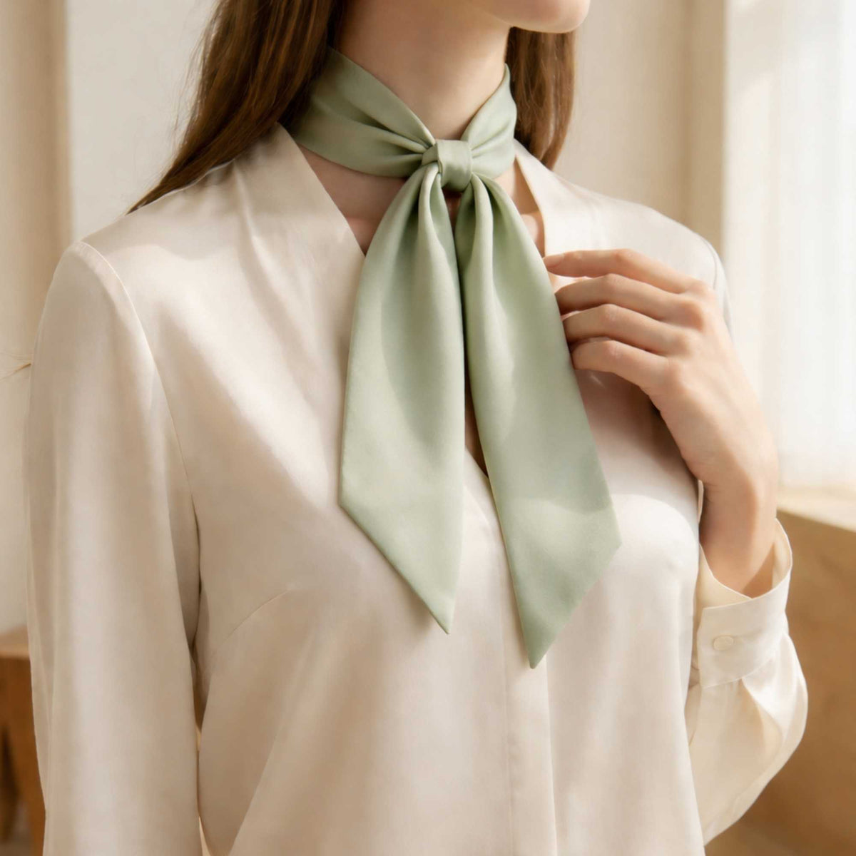 Person wearing a sage green skinny silk scarf tied around their neck with a neutral background