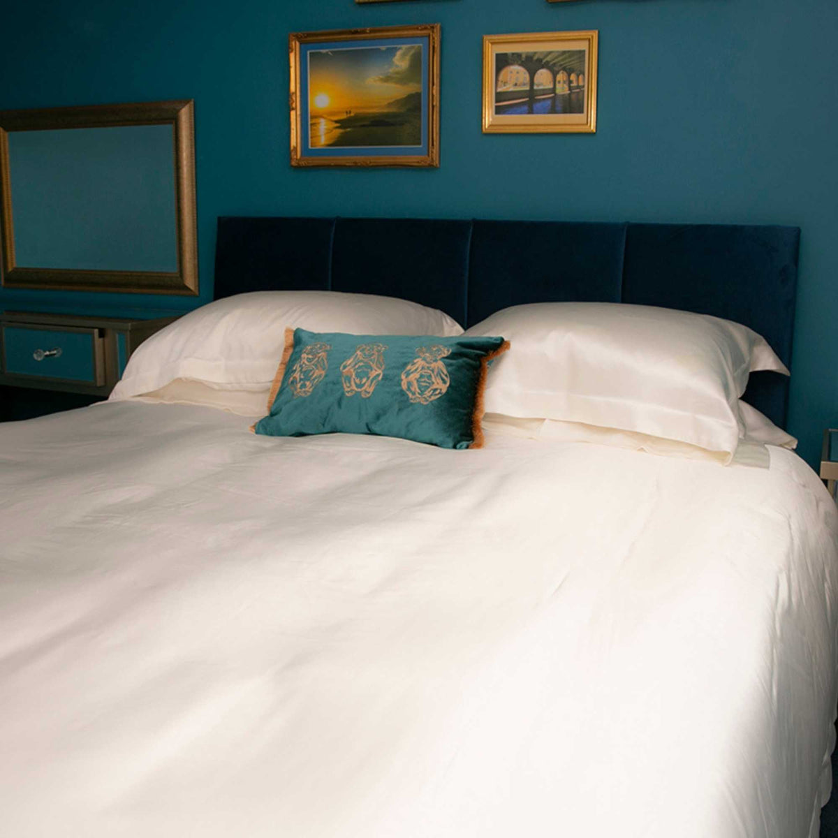 Bedroom with an ivory silk bed, pillows, and framed pictures on a blue wall.