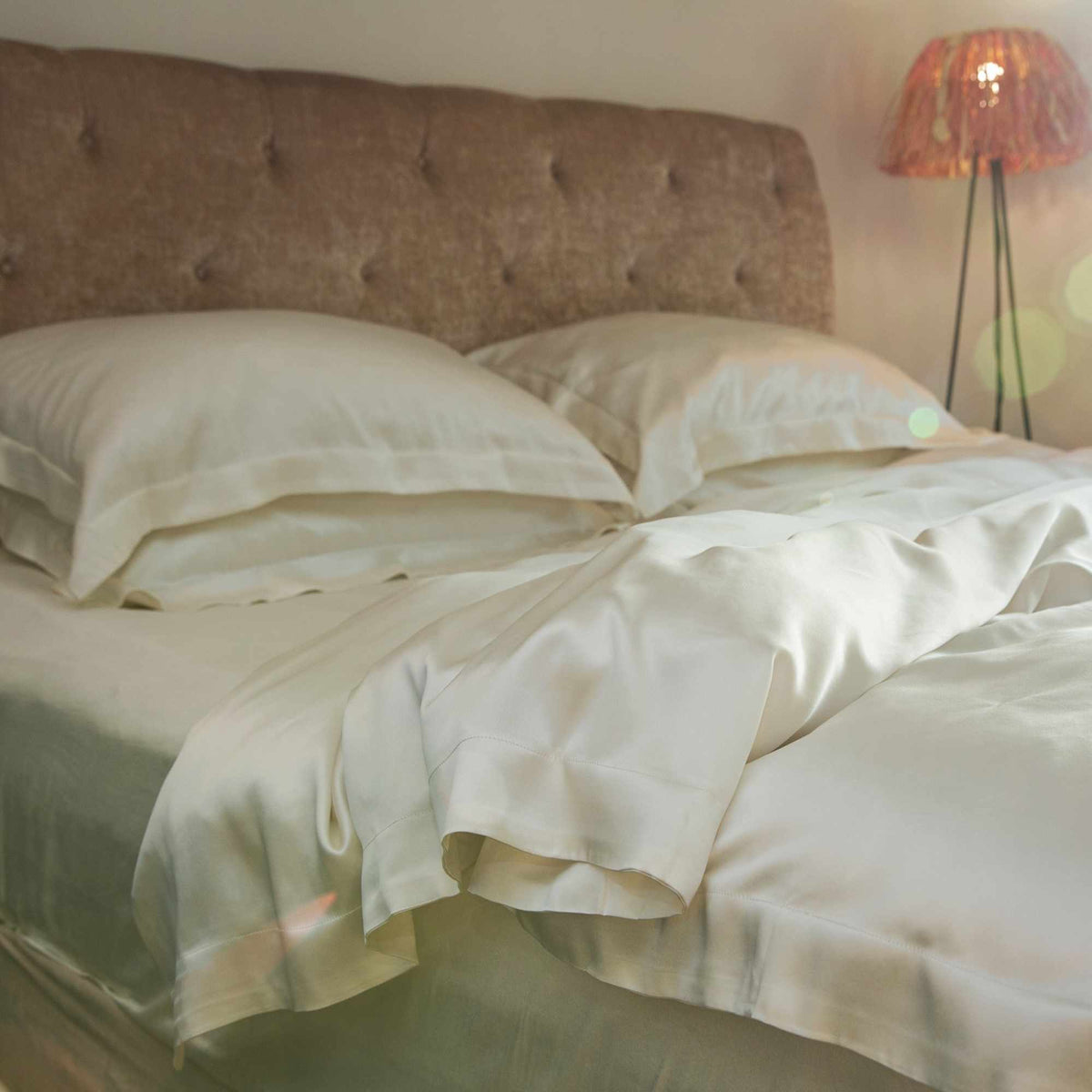 Neatly made bed with ivory silk bedding and pillows, illuminated by a lamp in a softly lit room.