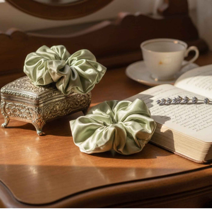 Two green oversized silk scrunchies on a wooden surface with a book and teacup in the background.