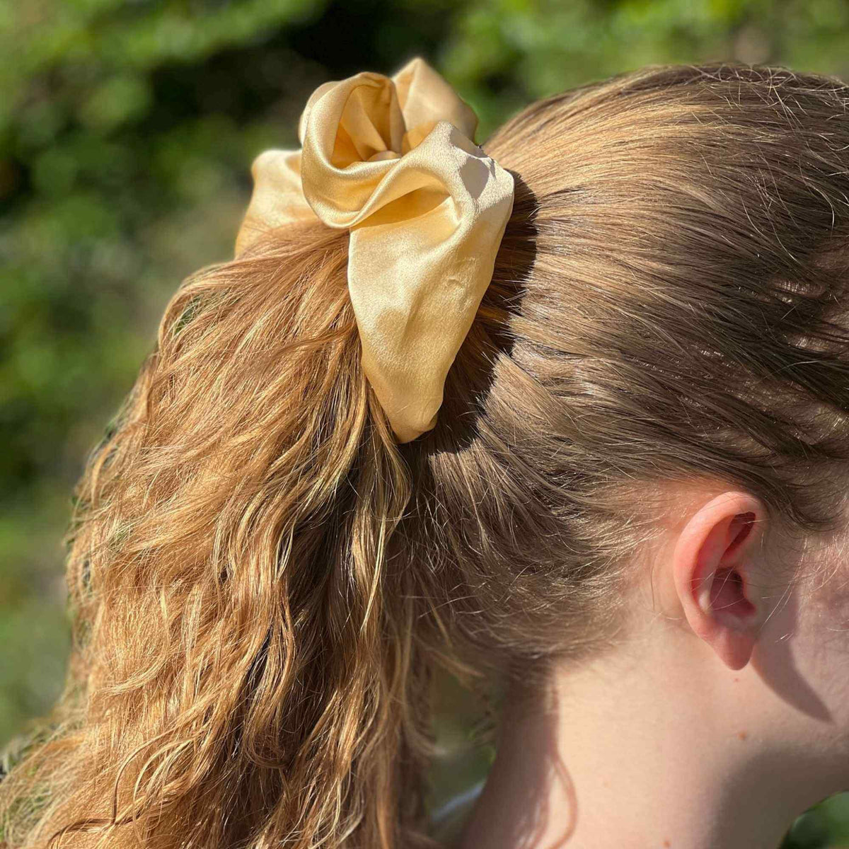 Person with a golden yellow oversized silk scrunchie in their hair against a blurred green background